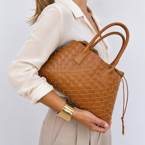 Può includere: Una borsa a mano color cuoio con un motivo in pelle intrecciata, due manici superiori arrotondati e un dettaglio con chiusura a coulisse sul lato. La borsa ha una forma rettangolare strutturata con una trama a griglia su tutta la superficie.