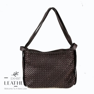 Op de afbeelding: Een donkerbruine leren schoudertas met een geweven patroon. De tas heeft een lange riem. De tekst "EXHIBIT LEATHER Welcome To A New Look" is zichtbaar.