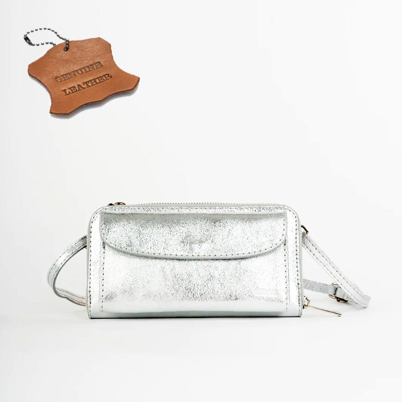 May include: A silver leather crossbody purse with a zipper closure and a small pocket on the front. The purse has a strap that can be adjusted to fit comfortably on the shoulder. The tag on the purse reads "Genuine Leather".
