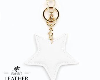 May include: A white leather star-shaped keychain with gold-colored hardware. The keychain has a gold chain and a clasp. The text "EXHIBIT LEATHER" and "Welcome To A New Look" are printed on the bottom.