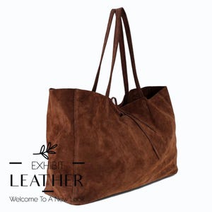 Op de afbeelding: Een grote, bruine suède tote bag met lange handvatten. De tas heeft een zachte, getextureerde uitstraling en een ruim interieur. De tekst "EXHIBIT LEATHER" en "Welcome To A New Look" zijn op de tas gedrukt.