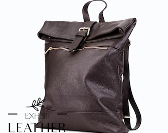Pode incluir: Uma mochila de couro castanho escuro com fecho de aba e fivela dourada. A mochila tem um bolso frontal com zíper e uma alça superior. O texto "EXHIBIT LEATHER" e "Welcome To A New Look" são visíveis.