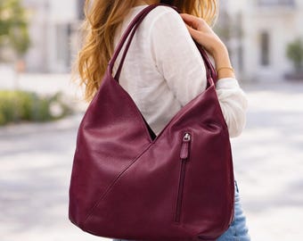 May include: A deep burgundy hobo bag with a textured surface, featuring a V-shaped front seam and a vertical zippered pocket on the side. The bag has two shoulder straps and a slouchy, unstructured silhouette that hangs from the shoulder.