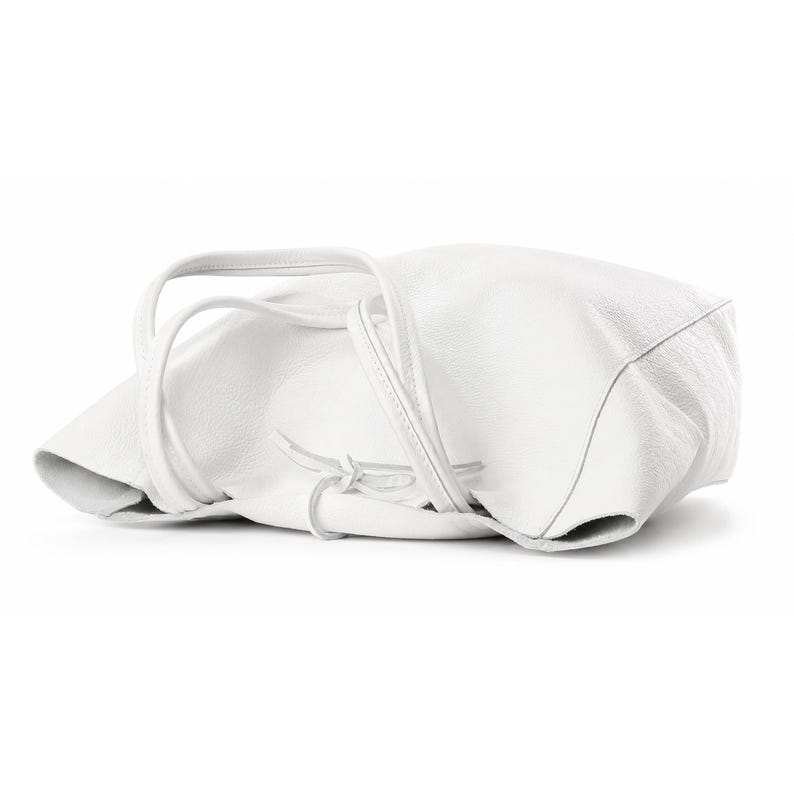 Pode incluir: Uma bolsa tote branca com superf&iacute;cie texturizada e duas al&ccedil;as finas de ombro. A bolsa apresenta um design macio e desestruturado com um pequeno detalhe de fecho de la&ccedil;o na parte frontal central, em uma cor branca brilhante.