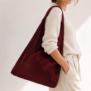 Op de afbeelding: Een diepbordeauxrode hobo-schoudertas met een zachte, matte textuur. De tas heeft een brede, geïntegreerde schouderband en een soepel, ongestructureerd silhouet dat op natuurlijke wijze langs de zijkant van het lichaam hangt.