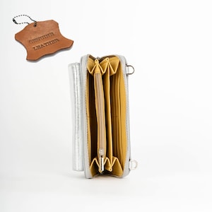 May include: A silver leather wallet with a zipper closure and multiple compartments. The wallet is open and the interior is lined with yellow fabric. The wallet has a small strap and a tag that reads "Genuine Leather".