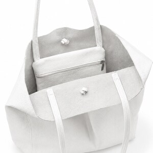 Pode incluir: Uma bolsa tote de couro branco com superf&iacute;cie texturizada, apresentando duas al&ccedil;as finas de ombro e uma bolsa com z&iacute;per branca correspondente fixada no interior. A bolsa tem um design aberto com um pequeno detalhe de n&oacute; na borda frontal.