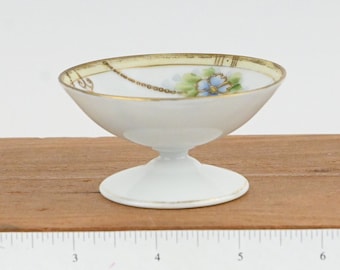 May include: A small white footed bowl with a gold rim and floral design. The bowl has a blue flower with green leaves and a gold chain design.