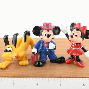 May include: A set of three plastic Disney character figurines. Mickey Mouse is wearing a blue suit and a red hat. Minnie Mouse is wearing a red dress with a white polka dot pattern and a red bow. Pluto is a yellow dog with black ears.