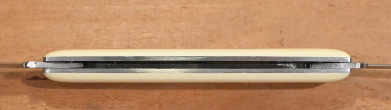 May include: A closed pocket knife with a cream-colored handle and a silver-colored blade. The knife is lying horizontally against a brown wooden surface. The blade is partially visible.