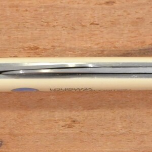May include: A closed pocket knife with a cream-colored handle and a silver blade. The word "LOUISIANA" is printed on the handle. The knife is lying on a wooden surface.