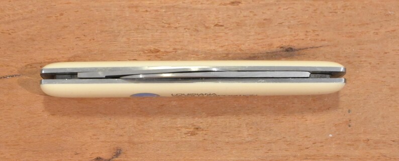 May include: A closed pocket knife with a cream-colored handle and a silver blade. The word "LOUISIANA" is printed on the handle. The knife is lying on a wooden surface.