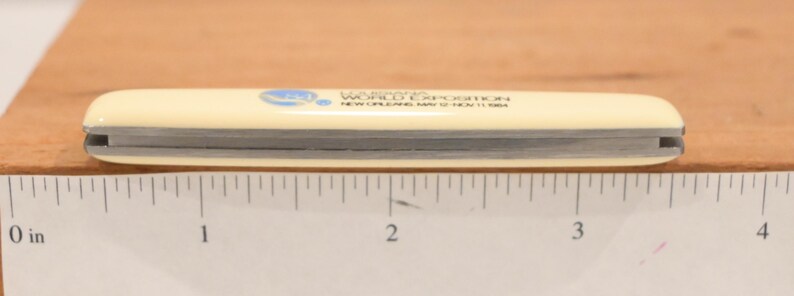 May include: A cream-colored pocket knife with a silver blade, measuring approximately 4 inches long. The knife has text that reads "Louisiana World Exposition New Orleans May 12-Nov 11 1984" and a blue logo.