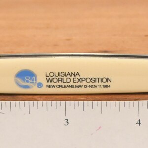 May include: A pocket knife with two blades extended. The handle is off-white with black trim and the text "LOUISIANA WORLD EXPOSITION NEW ORLEANS MAY 12 NOV 11 1984". The blades are silver and the knife is on a wooden surface.