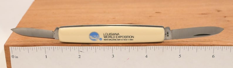 May include: A pocket knife with two blades extended. The handle is off-white with black trim and the text "LOUISIANA WORLD EXPOSITION NEW ORLEANS MAY 12 NOV 11 1984". The blades are silver and the knife is on a wooden surface.