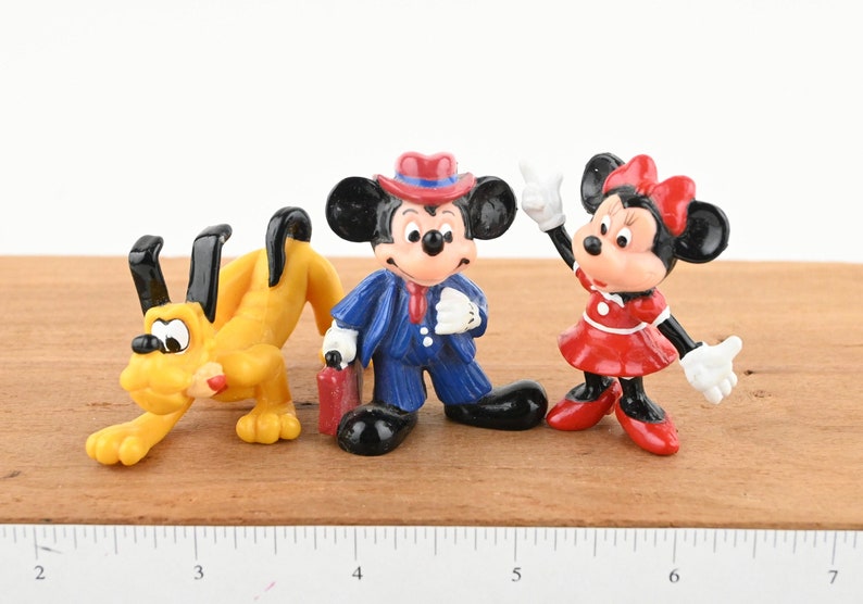 May include: A set of three plastic Disney character figurines. Mickey Mouse is wearing a blue suit and a red hat. Minnie Mouse is wearing a red dress with a white polka dot pattern and a red bow. Pluto is a yellow dog with black ears.