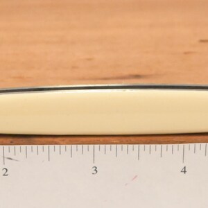 May include: A small, double-bladed pocket knife with a cream-colored handle and two stainless steel blades. The knife is open, displaying both blades. The handle is approximately 3 inches long. The knife is resting on a wooden surface with a ruler.