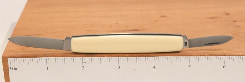 May include: A small, double-bladed pocket knife with a cream-colored handle and two stainless steel blades. The knife is open, displaying both blades. The handle is approximately 3 inches long. The knife is resting on a wooden surface with a ruler.