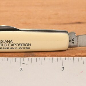 May include: A small, cream-colored pocket knife with a stainless steel blade. The knife's handle features the text "LOUISIANA WORLD EXPOSITION NEW ORLEANS MAY 12-NOV 11 1984" and a blue logo. The blade is partially open, revealing its sharp edge.