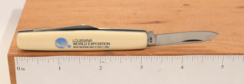 May include: A small, cream-colored pocket knife with a stainless steel blade. The knife's handle features the text "LOUISIANA WORLD EXPOSITION NEW ORLEANS MAY 12-NOV 11 1984" and a blue logo. The blade is partially open, revealing its sharp edge.