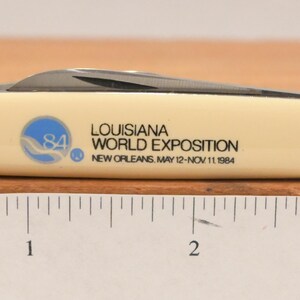 May include: A cream-colored pocket knife with a black blade. The knife features the text "LOUISIANA WORLD EXPOSITION NEW ORLEANS MAY 12-NOV 11 1984" and a blue and white logo. The knife is approximately 3 inches long.