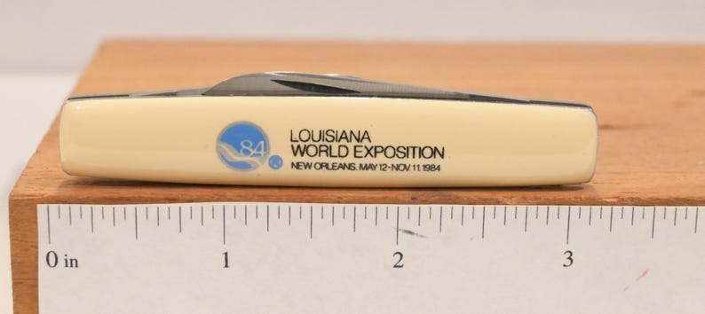 May include: A cream-colored pocket knife with a black blade. The knife features the text "LOUISIANA WORLD EXPOSITION NEW ORLEANS MAY 12-NOV 11 1984" and a blue and white logo. The knife is approximately 3 inches long.