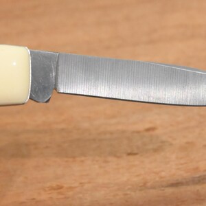 May include: Close-up of a small, folding knife with a cream-colored handle and a silver blade. The blade is partially open, revealing its sharp edge. The knife rests on a wooden surface.
