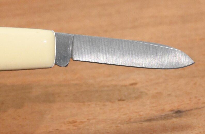 May include: Close-up of a small, folding knife with a cream-colored handle and a silver blade. The blade is partially open, revealing its sharp edge. The knife rests on a wooden surface.