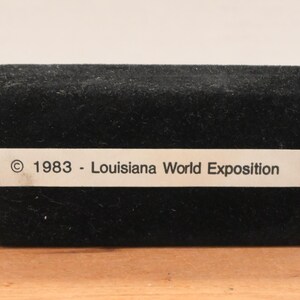 May include: A black velvet box with a rectangular shape. A white label on the front reads "&copy; 1983 - Louisiana World Exposition". The box is set on a wooden surface.