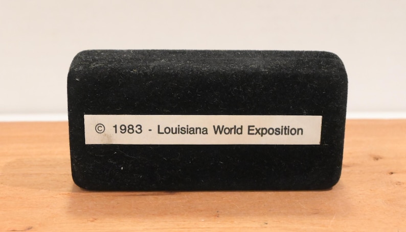 May include: A black velvet box with a rectangular shape. A white label on the front reads "&copy; 1983 - Louisiana World Exposition". The box is set on a wooden surface.
