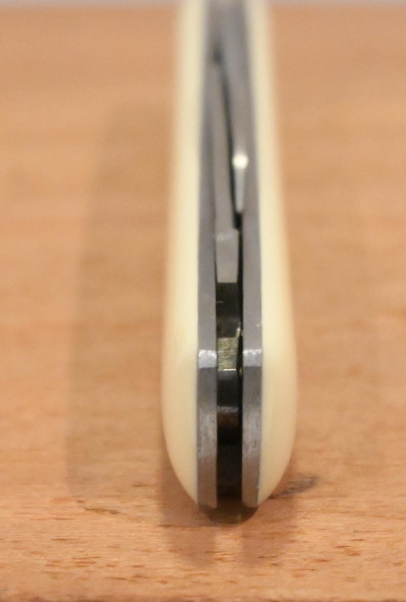May include: Close-up of a pocket knife with a cream-colored handle and a silver blade. The knife is closed, showing the handle's smooth, rounded shape and the metal components. The blade is partially visible.