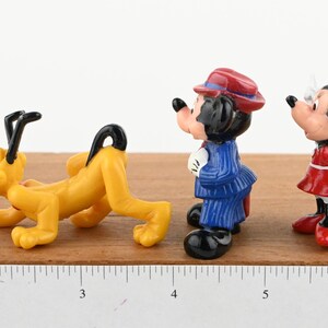 May include: A set of three small plastic figurines of Disney characters. The figurines are of Pluto, Mickey Mouse, and Minnie Mouse. Pluto is yellow and is in a crawling position. Mickey Mouse is wearing a blue suit and a red hat. Minnie Mouse is wearing a red dress with white polka dots and a red bow. The figurines are on a wooden surface with a ruler for scale.