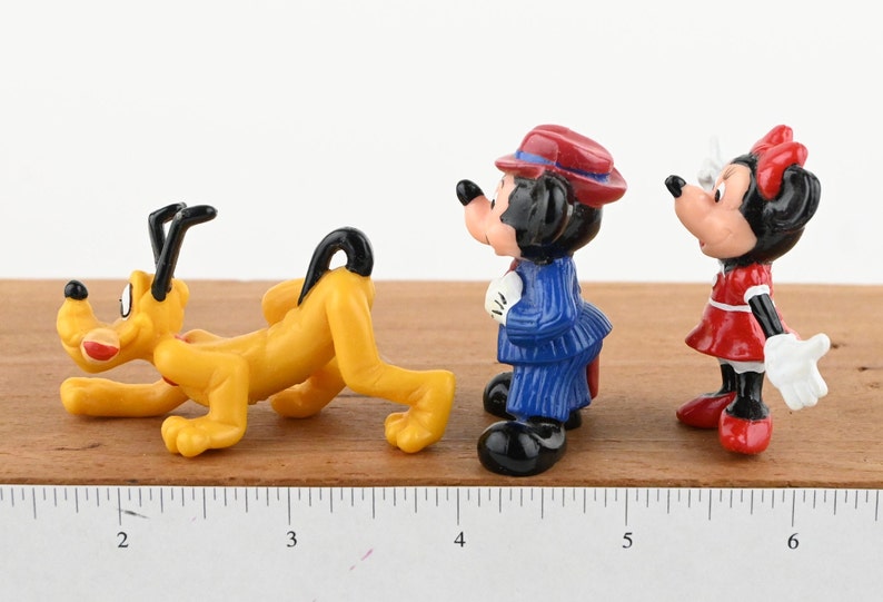 May include: A set of three small plastic figurines of Disney characters. The figurines are of Pluto, Mickey Mouse, and Minnie Mouse. Pluto is yellow and is in a crawling position. Mickey Mouse is wearing a blue suit and a red hat. Minnie Mouse is wearing a red dress with white polka dots and a red bow. The figurines are on a wooden surface with a ruler for scale.