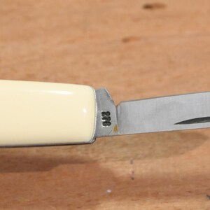 May include: A small, folding pocket knife with a cream-colored handle and a stainless steel blade. The blade is partially open, revealing a sharp edge. The handle has a smooth, glossy finish. The blade has the number "276" etched on it.