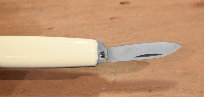 May include: A small, folding pocket knife with a cream-colored handle and a stainless steel blade. The blade is partially open, revealing a sharp edge. The handle has a smooth, glossy finish. The blade has the number "276" etched on it.