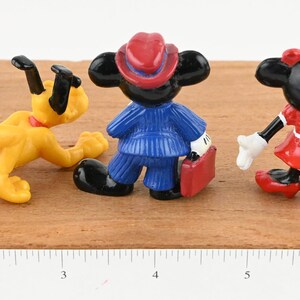 May include: Three small plastic figurines of Disney characters: Pluto the dog in yellow, Mickey Mouse in a blue suit and red hat, and Minnie Mouse in a red dress. The figurines are posed in a standing position.