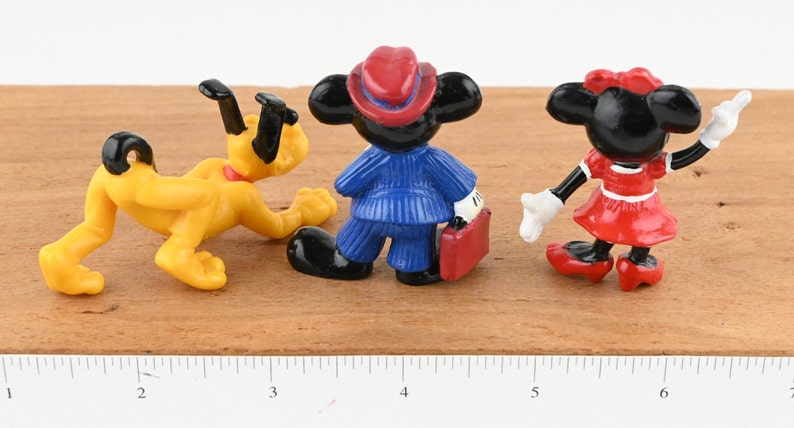 May include: Three small plastic figurines of Disney characters: Pluto the dog in yellow, Mickey Mouse in a blue suit and red hat, and Minnie Mouse in a red dress. The figurines are posed in a standing position.