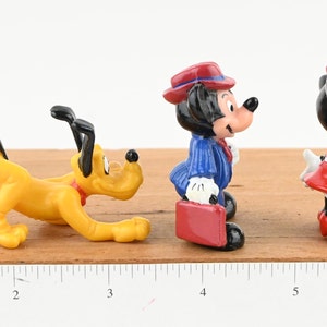May include: A set of three small plastic figurines of Disney characters. The figurines are of Pluto, Mickey Mouse, and Minnie Mouse. Pluto is yellow and is in a crouching position. Mickey Mouse is wearing a blue suit and a red hat. Minnie Mouse is wearing a red dress and a red bow. The figurines are on a wooden surface with a ruler.