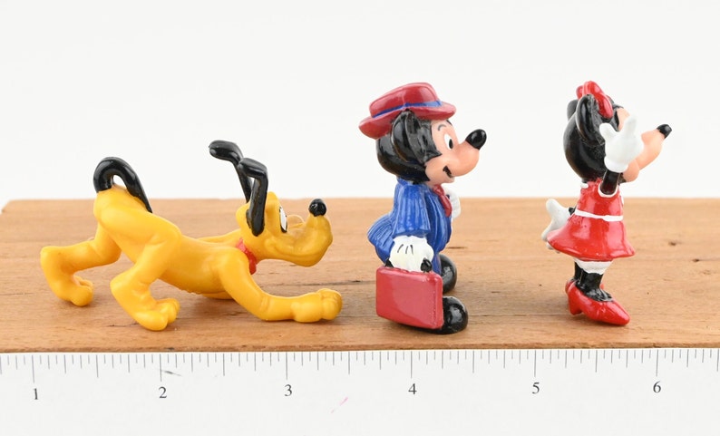May include: A set of three small plastic figurines of Disney characters. The figurines are of Pluto, Mickey Mouse, and Minnie Mouse. Pluto is yellow and is in a crouching position. Mickey Mouse is wearing a blue suit and a red hat. Minnie Mouse is wearing a red dress and a red bow. The figurines are on a wooden surface with a ruler.