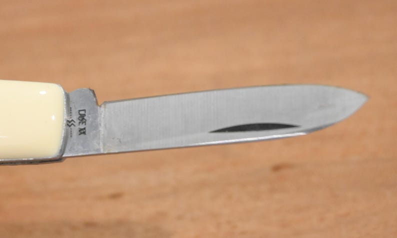 May include: Close-up of a stainless steel pocket knife blade with a cream-colored handle. The blade has a curved edge and a small indentation. The knife is marked with the text "CASE XX".
