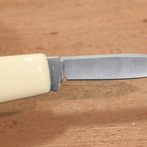 May include: A small, folding knife with a cream-colored handle and a silver blade. The handle features a blue and white emblem. The blade is open, revealing its sharp edge. The knife is designed for everyday use.