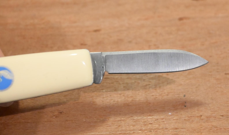 May include: A small, folding knife with a cream-colored handle and a silver blade. The handle features a blue and white emblem. The blade is open, revealing its sharp edge. The knife is designed for everyday use.