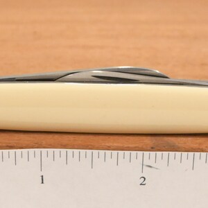 May include: A closed pocket knife with a cream-colored handle and a silver blade. The knife is approximately 3 inches long. The knife is on a wooden surface.