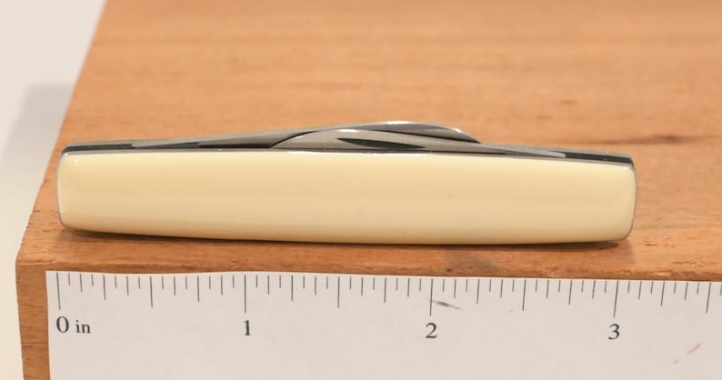 May include: A closed pocket knife with a cream-colored handle and a silver blade. The knife is approximately 3 inches long. The knife is on a wooden surface.