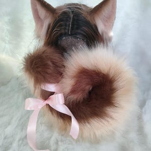 Op de afbeelding: Een bruine en witte nep bontstaart met een roze strik eromheen gebonden. De staart is bevestigd aan een kattenkostuum.