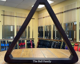 Puede incluir: Un estante de triángulo de billar de madera marrón oscuro con el texto "The Ball Family" grabado en la parte inferior.