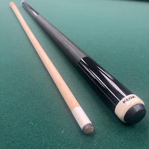 May include: A two-piece pool cue featuring a light-coloured wooden shaft with a white ferrule and a black butt section. The butt end is finished with a white collar displaying the black text KODA and a black rubber bumper at the very base.