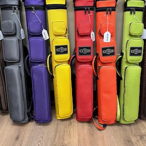 May include: A row of six pool cue cases in grey, purple, yellow, red, orange, and lime green. Each case features two external zippered pockets, a top handle, and a black rectangular patch with the white text C.P. DEAN printed on the lower pocket.