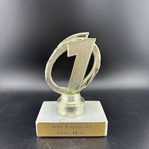 May include: A gold-colored trophy featuring a large number one centered within an oval frame. The trophy is mounted on a white rectangular base with a gold-toned metal plate at the front that displays the text Your Engraving Goes Here.