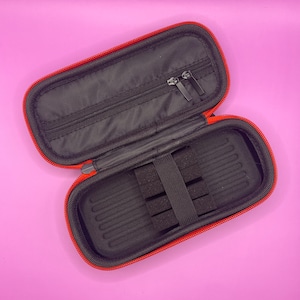 May include: Red zippered case with black interior and a zippered pocket. The case has a black foam insert with slots for holding multiple items.